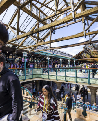 Busy trade show visitors moving through a historic UK exhibition venue with wrought-iron architecture and a packed upper gallery.