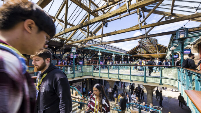Busy trade show visitors moving through a historic UK exhibition venue with wrought-iron architecture and a packed upper gallery.