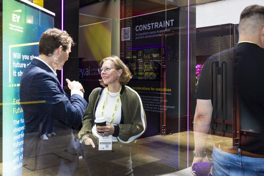 Two attendees having a conversation at an exhibition stand, with EY branded signage and digital displays in the background