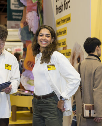 Exhibition stand staff in white branded polo shirts chatting and reviewing a tablet in front of a yellow 'Fresh for...' trade show booth