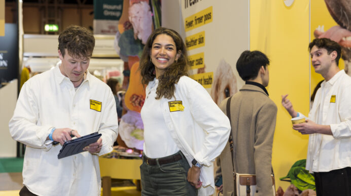 Exhibition stand staff in white branded polo shirts chatting and reviewing a tablet in front of a yellow 'Fresh for...' trade show booth