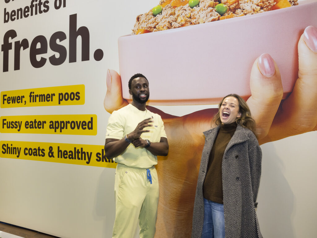 Exhibition stand staff member mid-conversation with a laughing visitor in front of a fresh dog food brand's benefits wall.