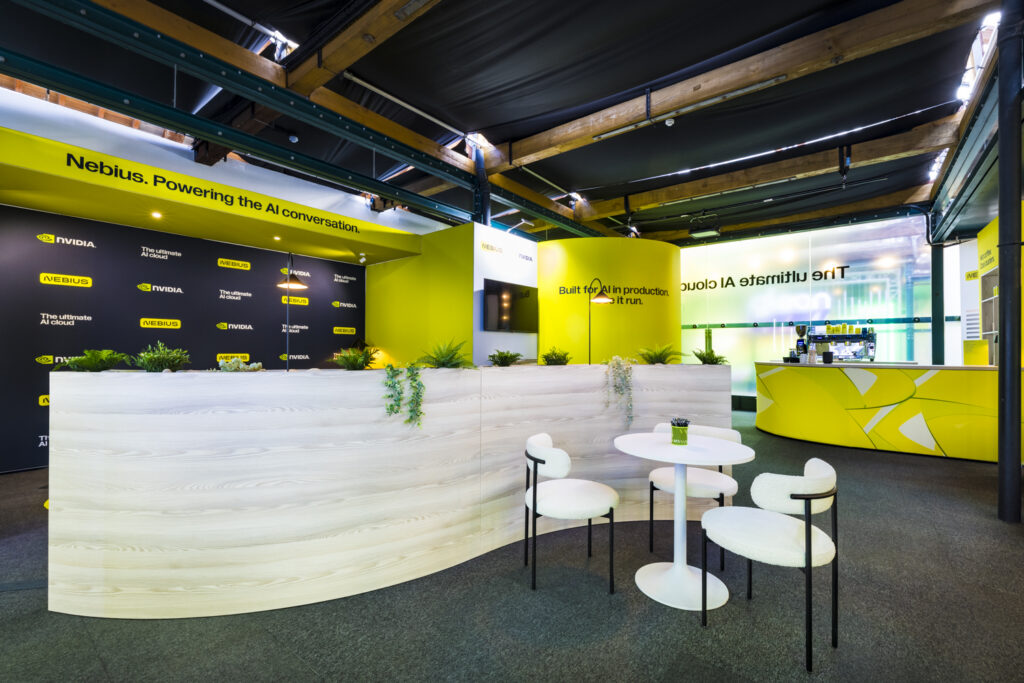 Nebius exhibition stand with curved white reception desk, greenery accents, black and yellow branded messaging walls, and white round meeting tables with white and black chairs