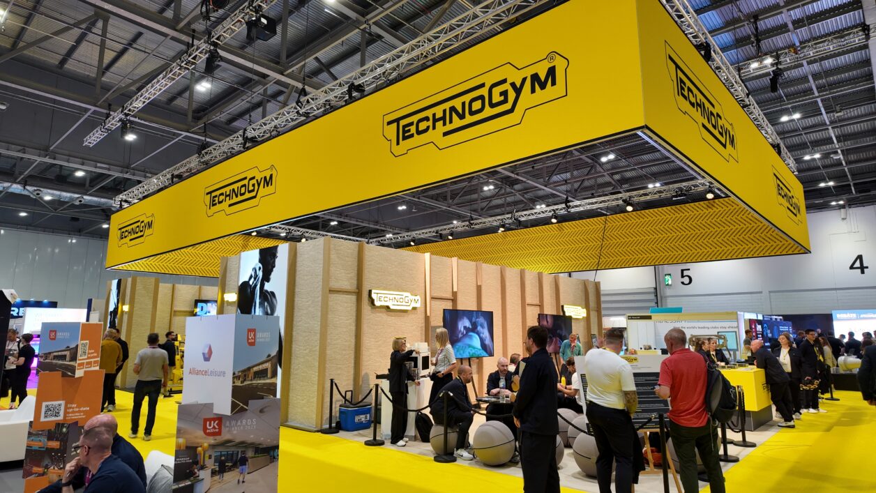 Technogym exhibition stand with bold yellow branding and overhead signage on a busy trade show floor.