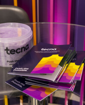 Alt text: Branded Tecna candy floss tub and "Push the Possible" brochures on a glass table at Confex 2026, set against a colourful backlit exhibition stand
