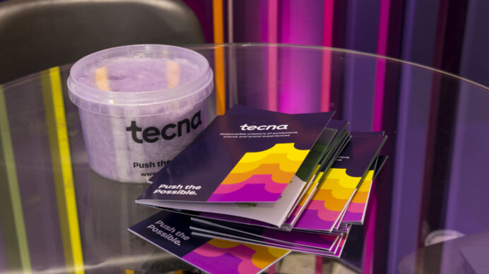 Alt text: Branded Tecna candy floss tub and "Push the Possible" brochures on a glass table at Confex 2026, set against a colourful backlit exhibition stand