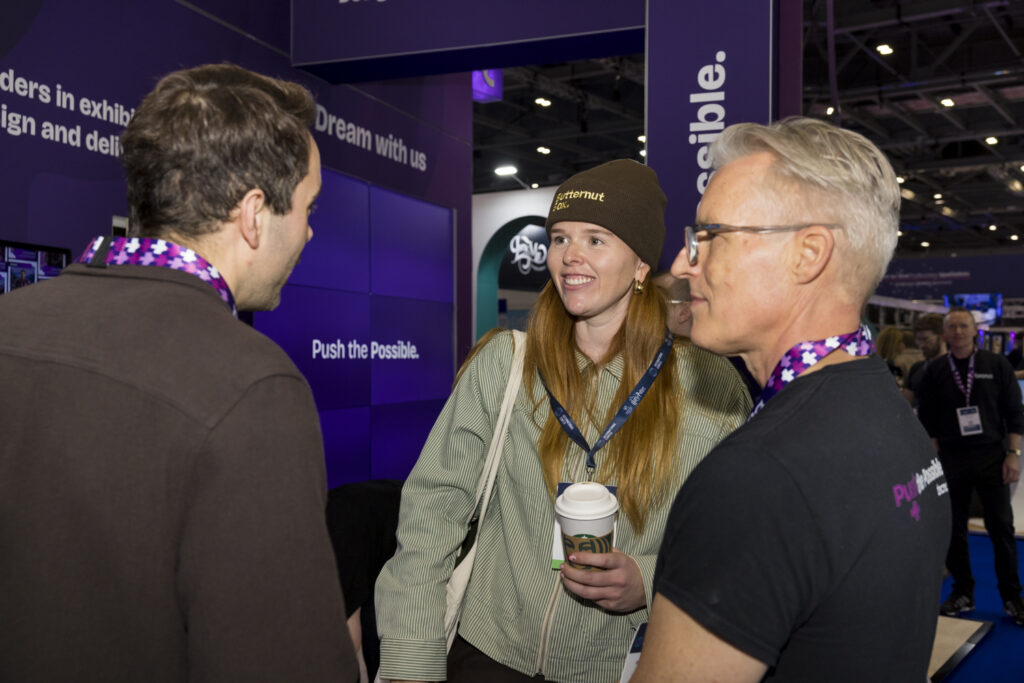 Tecna stand staff member talking with two visitors at a trade show booth branded with 'Push the Possible