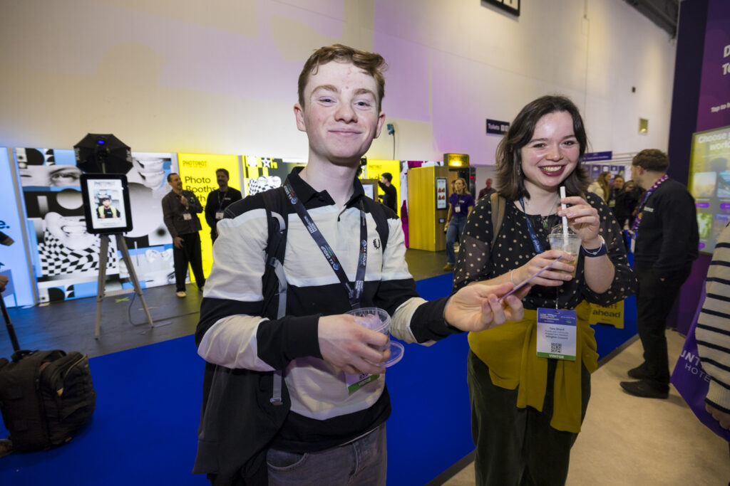 Two exhibition attendees smiling and holding drinks at a trade show, with exhibitor booths and a crowd visible in the background